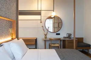 A modern minimalist bedroom with wooden furniture and white bedding, featuring a round mirror, a small table, and a comfortab