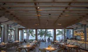 Elegant coastal restaurant interior with wooden tables, chairs, and glass windows showcasing a scenic ocean view.