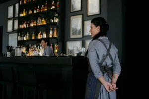 A woman wearing an apron standing at a bar with a backdrop of shelves filled with bottles and framed pictures on the wall.