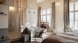A minimalist bathroom featuring a white and marble floor with a walk-in shower and a sunlit room with a desk and chair.