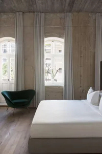 A minimalist bedroom featuring a white bed with white linens, a green armchair, and white curtains.
