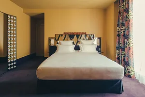 A spacious modern bedroom featuring a bed with white linens, a geometric headboard, and colorful curtains against a mustard y