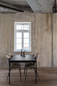 A modern, minimalist dining area with concrete walls, a black dining table, beige chairs, and tall candle holders, illuminate