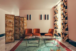 A modern living room with orange upholstered chairs, a glass coffee table, geometric-patterned curtains, and marble accents.