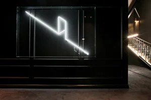 A minimalist installation art piece featuring a neon light sculpture mounted on a black wall.