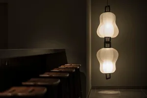 A row of stools in a dimly lit restaurant bar with a minimalist design, featuring a decorative paper lamp hanging from the ce