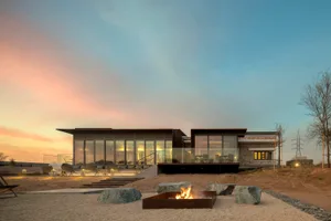 Modern minimalist house with large glass windows and a stone-clad exterior, featuring a fire pit on the patio.