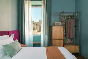 Modern hotel room with neutral bed and furniture, blue walls, and a view outside through sheer curtains.
