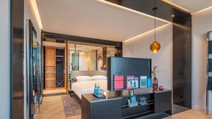 Modern hotel room with wooden flooring and glass walls, featuring a bed, nightstand, and magazines.