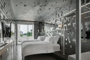 Modern minimalist hotel room with metallic mosaic tiles, bed with white bedding, and floor-to-ceiling windows.