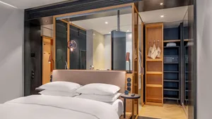 A modern bedroom featuring a large bed with white sheets, a bedside table with a camera, and a wooden floor.