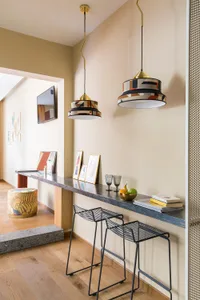 Modern kitchen with marble countertops, colorful pendant lights, and black stools. Wooden floor and framed art on the wall.