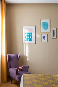 A purple upholstered wingback chair in a well-lit bedroom with a yellow curtain, abstract art wall hanging, and a bed with a 