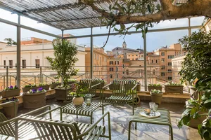 A rooftop patio with metal chairs and tables surrounded by potted plants, set against a backdrop of buildings and a church sp