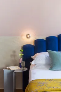 Modern bedroom with a blue upholstered bed, concrete walls, and a glass vase with a green plant.