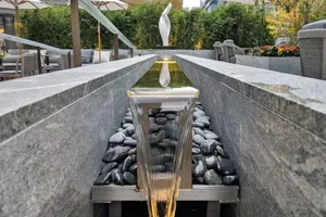 A minimalist water feature consisting of granite, stainless-steel, and glass materials, with a reflective glass element and s