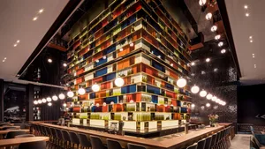 A modern restaurant interior with wooden tables and chairs, colorful shelves, and hanging lights.