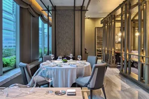 Modern restaurant dining room with marble flooring, metal railings, and floor-to-ceiling windows overlooking cityscape.