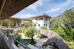 White stucco villa with a pool and lush greenery, featuring a thatched roof cabana and wooden patio.