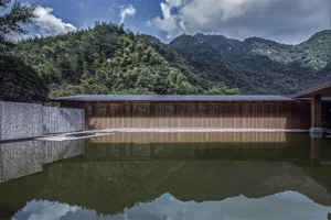 A minimalist landscape featuring a wooden building reflected in a calm pool of water, with dense green foliage and mountains 