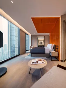 Modern hotel room with a bed, coffee table, and open city view through large windows.