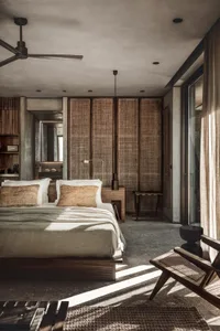 Minimalist bedroom with concrete walls and floor, featuring a bed with white bedding and beige cushions, a wooden chair, and