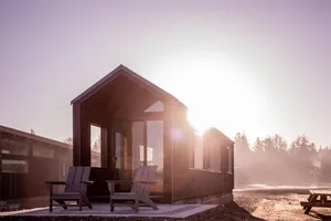 Sunlit wooden shed with Adirondack chairs in front, surrounded by trees.