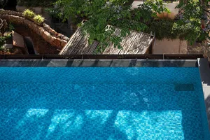 A modern poolside patio with blue tiled walls and a wooden roof structure surrounded by greenery.