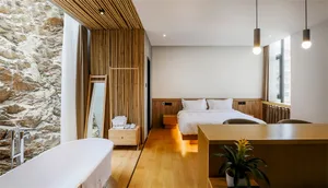 Modern minimalist bedroom featuring a wooden bed and floor, with natural light streaming through a large window and a stone w