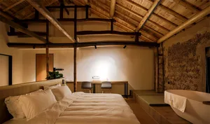 A minimalist bedroom featuring wooden beams on the ceiling, a bed with white bedding, and a small desk area.