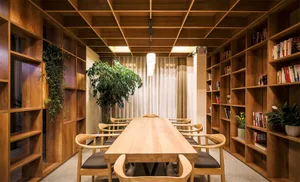 A modern library or study room with wooden furniture and shelves, featuring a wooden table, chairs, and potted plants.