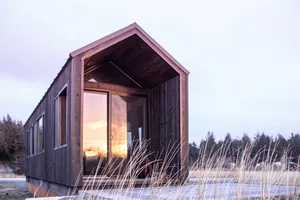 A modern, minimalist wooden house with large windows reflecting a sunset.