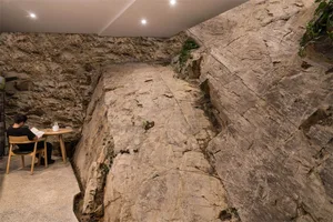 A person sits at a wooden table and reads a book in a cave-like interior space with stone walls and a ceiling light.