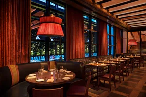 A luxurious dining room with red velvet curtains and wooden flooring, featuring round tables set for dinner with wine glasses