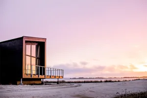 Minimalist, modern cabin with large windows and wooden exterior on a sandy beach at sunset.