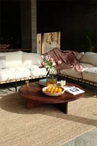 A modern living room featuring a round wooden table with a bowl of oranges, a vase of flowers, and various other items.