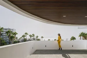 A woman in a yellow suit standing on a modern balcony with steel and white concrete elements, overlooking a coastal view with