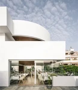 White minimalist restaurant with glass walls and white furniture under a cloudy sky.