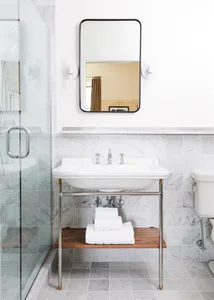 A minimalist bathroom with a white porcelain sink, marble tiles, and a wooden shelf with folded white towels beneath it.