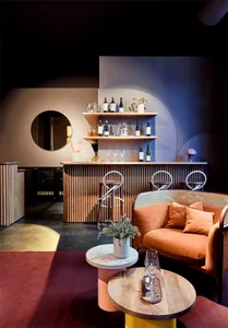 Modern bar area with wooden counter, shelves, orange couch, and round tables.