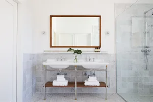 A modern bathroom with white marble tile walls, floor, and two white sinks with chrome faucets and a wooden shelf below. A gr