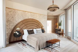 A minimalist bedroom featuring a bed with a textured wall fabric headboard, white bedding, and a wooden bench.