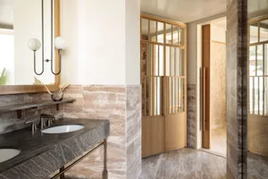 A minimalist bathroom with stone tiles and a double sink, featuring a wooden door and a mirror above the sink.