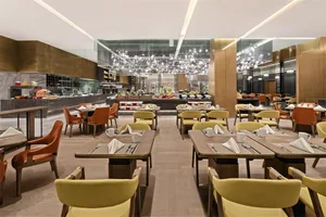 A modern restaurant interior featuring tables and chairs with wooden tops, orange and yellow upholstered chairs, and metallic