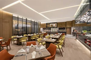A spacious, modern restaurant interior with wooden tables, yellow chairs, and a well-lit kitchen area.