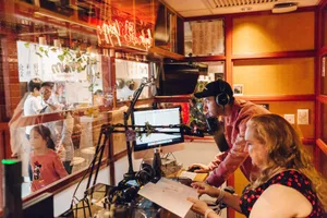 兩位工作人員在電台工作室內操作電腦螢幕、麥克風與文件。
