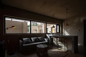 A minimalist indoor residential space with concrete walls and floor, featuring large windows, a sofa, and a bar counter.