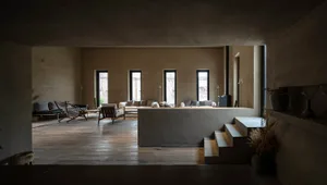 A minimalist living room featuring concrete walls and floors, with a central raised seating area and large windows.