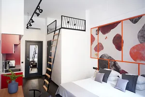 A modern minimalist bedroom with a bed, pillows, and abstract wall art. A loft area with a ladder staircase leads to another 