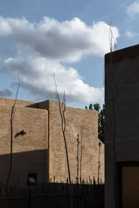 A sandstone building with thin trees and a wooden fence against a cloudy sky.